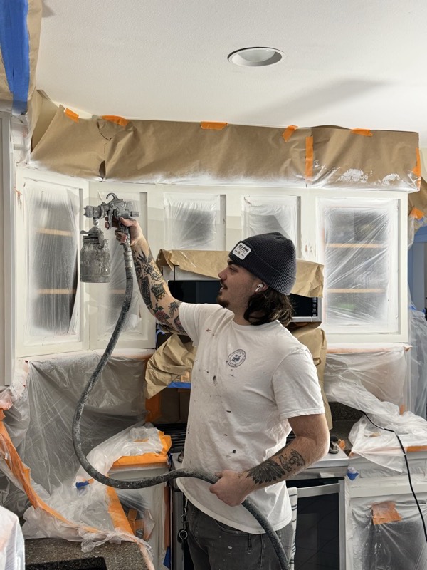 Painter spray-finishing cabinets in a fully masked kitchen
