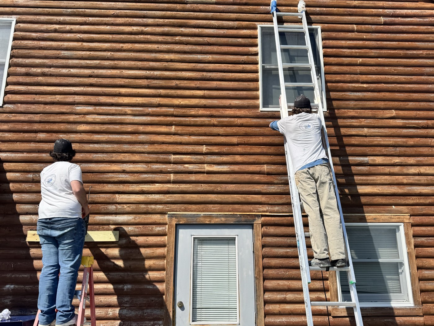 Crew staining the log walls of a log home from a ladder and step stool