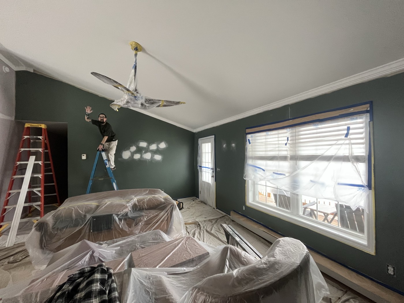Crew patching drywall from a ladder in a vaulted room with the furniture and floor covered