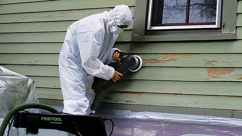 Worker in a protective suit and respirator sanding siding with a HEPA dust extractor and plastic sheeting below